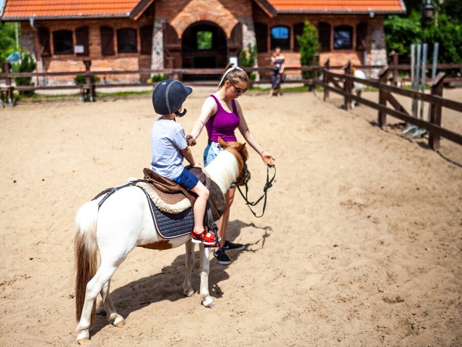 Child wearing a helmet on a small pony led by an instructor in a sandy riding arena beside a rustic stable.