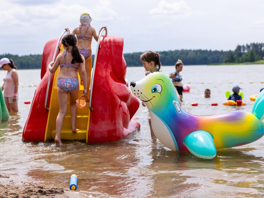 Children on a red waterslide at a shallow lake beach beside a colorful inflatable seal in family-friendly lakeside play area.