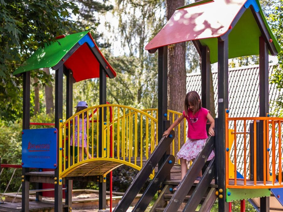 Family-friendly hotel playground with children on colorful climbing structure, bridge and shaded roofs.