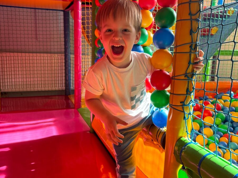 Laughing young boy climbing into a colorful indoor soft-play ball pit in a family-friendly hotel's kids' playroom.
