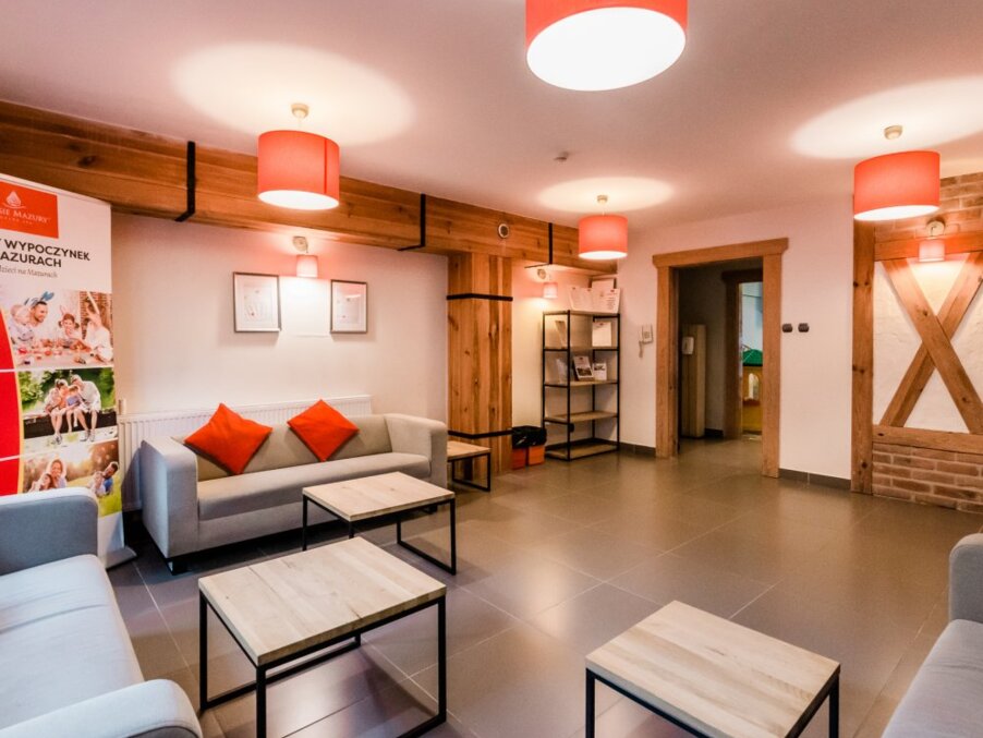 Warm hotel lounge with gray sofas, orange cushions, wood beams, pendant lights, coffee tables and a brochure rack.