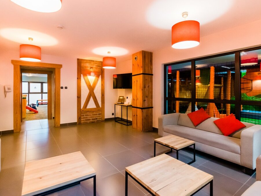 Family hotel lounge with sofa and red cushions, wooden tables, warm pendant lights and view into colorful indoor play area.