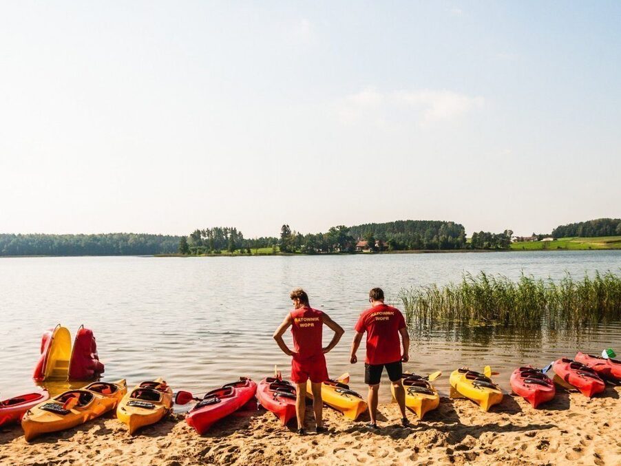 Wypożyczalnia kajaków nad jeziorem: czerwone i żółte kajaki ustawione na piasku, dwaj ratownicy przygotowują sprzęt nad spokojną wodą