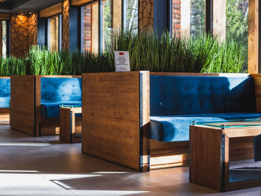 Hotel lounge booths with deep blue velvet seats, wooden dividers, glass tables and tall greenery by sunlit windows