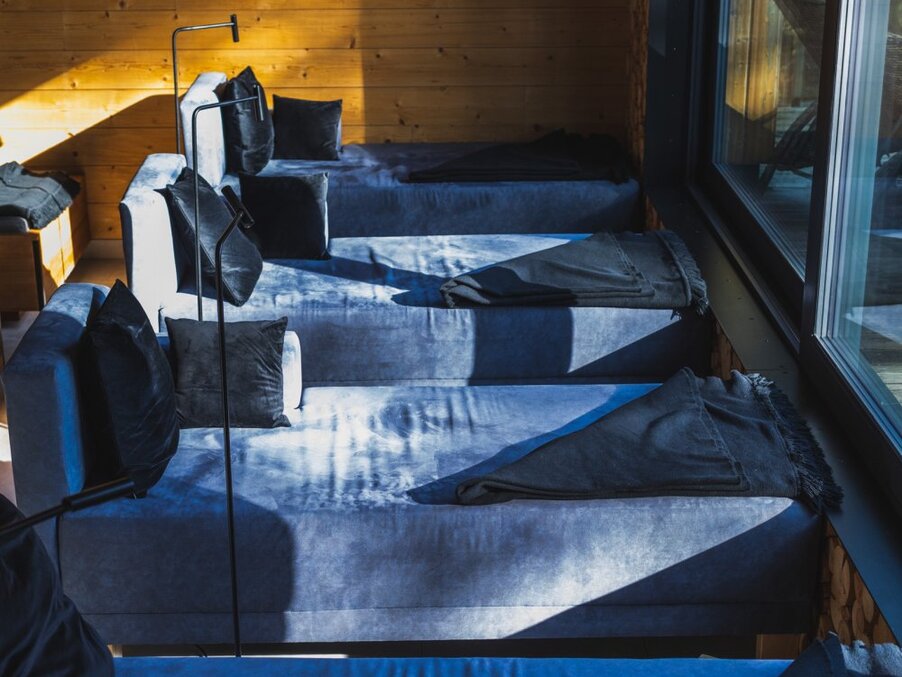 Sunlit row of blue velvet daybeds with black throws and cushions in a wood-paneled relaxation lounge by a large window