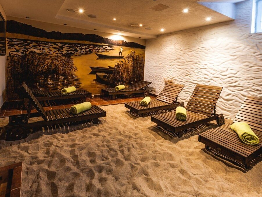 Indoor spa relaxation room with sand floor, wooden lounge chairs each with rolled green towel, warm mural of boats and reeds.