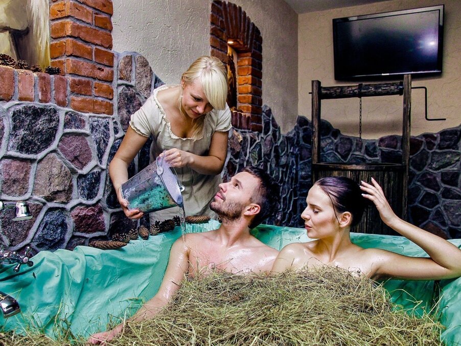 Couple relaxing in a hay-filled spa tub as attendant pours water from a metal bucket in a rustic stone and brick treatment room.