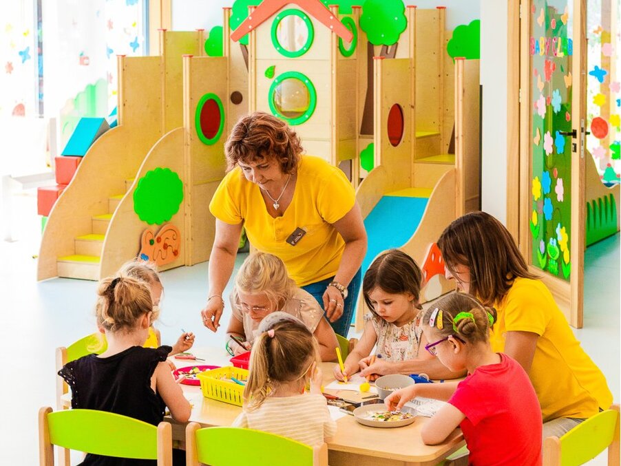 Hotel kids' club craft session with two staff supervising children at a table, colorful playhouse and indoor slides behind