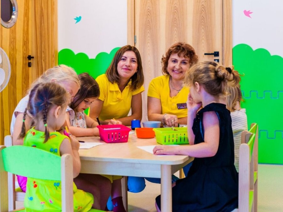 Children's activity table in hotel kids' club — two staff in yellow shirts supervising arts-and-crafts with five guests.