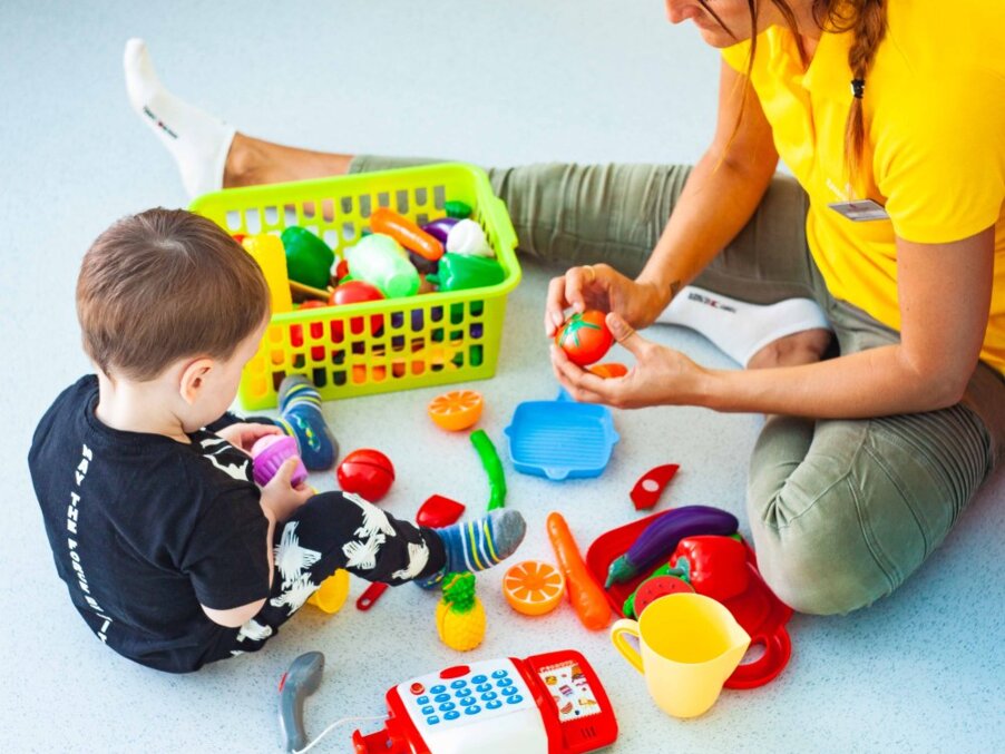 Young child and staff member sitting on the floor playing with colorful toy fruits and a play cash register — kids' club activity.