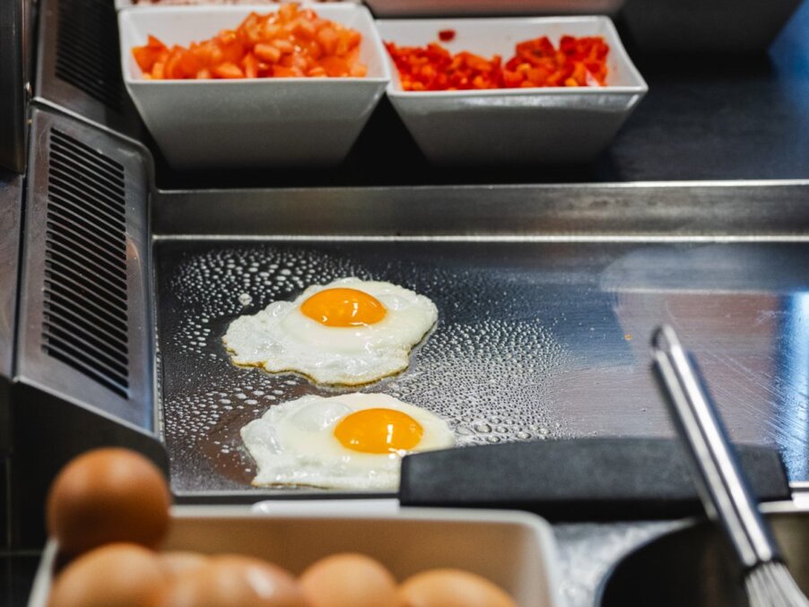 Live breakfast station with two sunny-side-up eggs on a hot stainless-steel griddle, chopped tomatoes and peppers in bowls nearby.