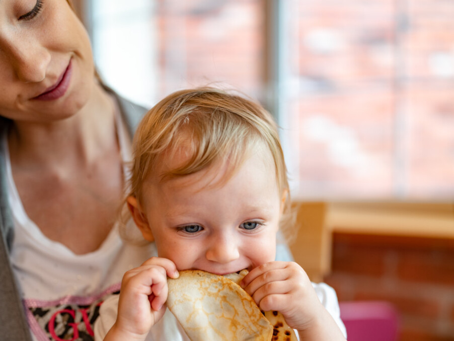 Toddler on mother's lap biting a warm crepe during hotel breakfast in a cozy, family-friendly dining area.