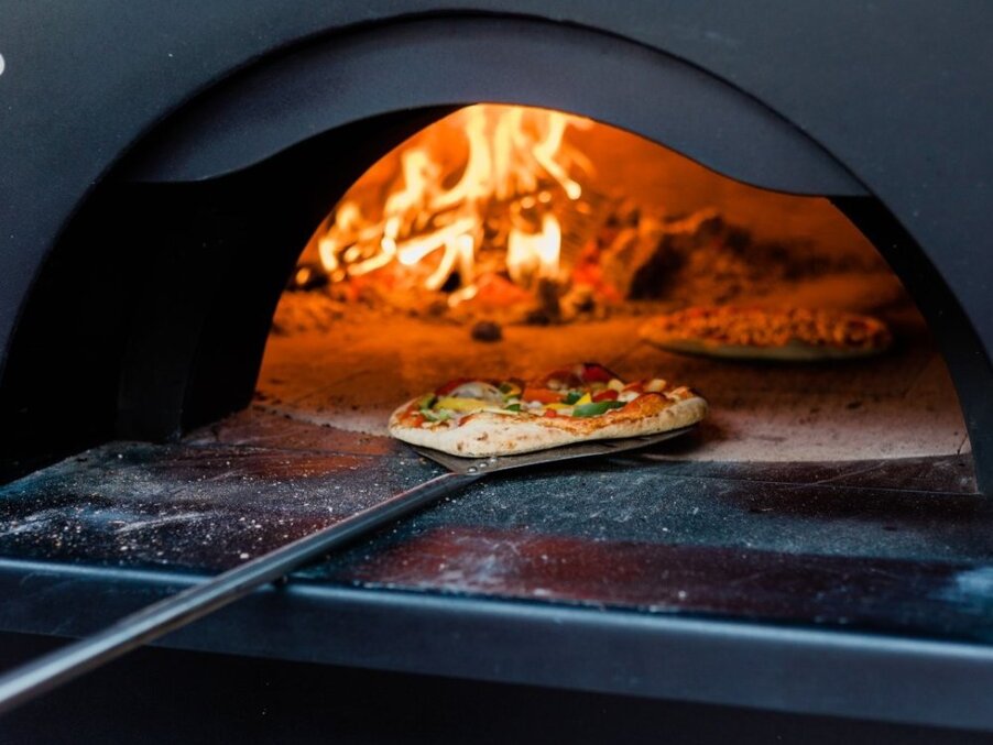 Wood-fired pizza on a peel inside a glowing stone oven, bubbling cheese, charred crust and fresh vegetable toppings