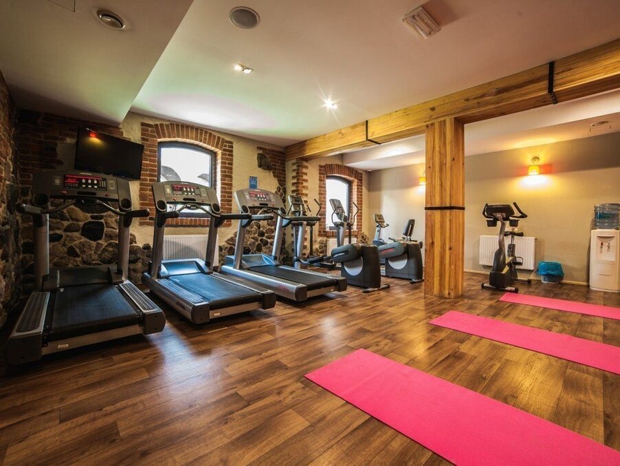 Hotel gym with treadmills, stationary bikes and pink yoga mats on warm wood floor, brick and stone accents, arched windows.