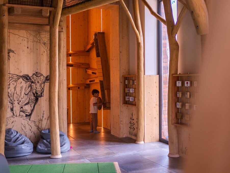Indoor family playroom with natural wood 'tree' structures, cow mural, bean bags, child playing with interactive wooden games by window light.