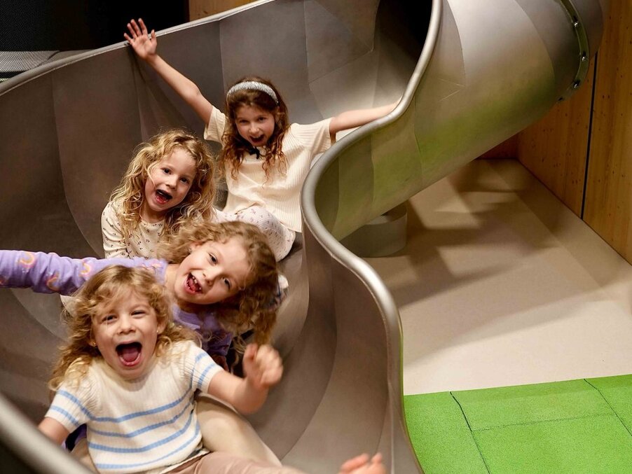 Four joyful children sliding down a wide indoor metal slide in the hotel's family playroom