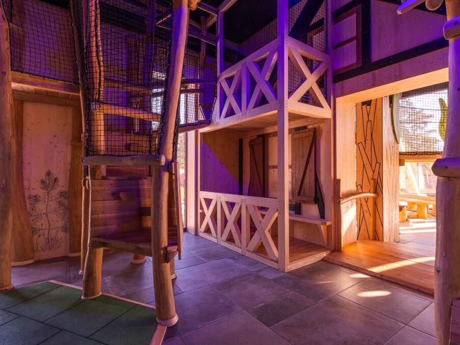 Hotel's rustic indoor kids' playroom with wooden climbing frames, net tunnels, mezzanine lofts and warm purple-accent lighting, family-friendly.