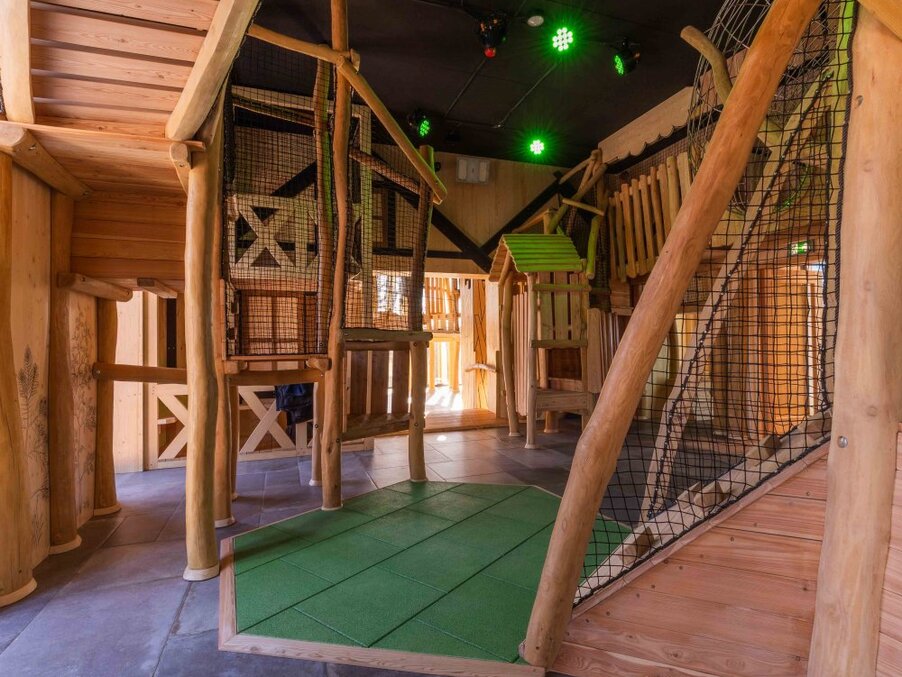 Indoor children's play area with rustic wooden treehouse frames, climbing nets, soft green safety mat and ceiling lights.