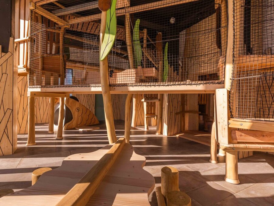 Indoor kids' play area with multi-level wooden climbing structure, safety netting and fish-shaped slide in warm sunlight