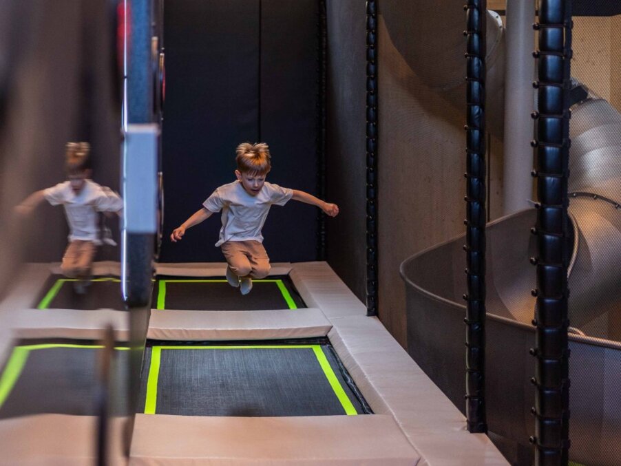 Child jumping on padded indoor hotel trampolines, mirrored wall reflection beside a spiral slide in the family play area.