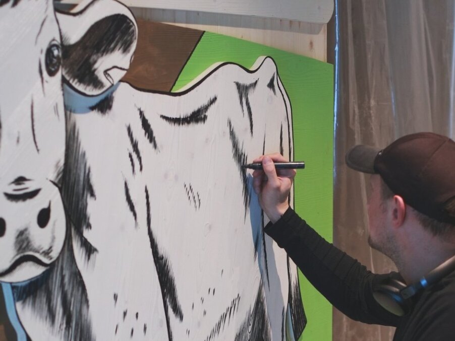 Artist wearing cap and headphones uses marker to draw black-and-white cow mural on wooden panel with green background