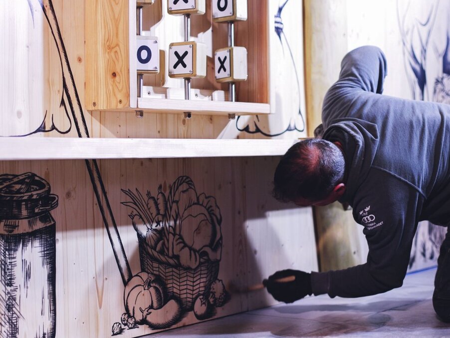 Person crouched painting a black hand-drawn vegetable basket mural on a light wooden panel under a shelf with tic-tac-toe blocks