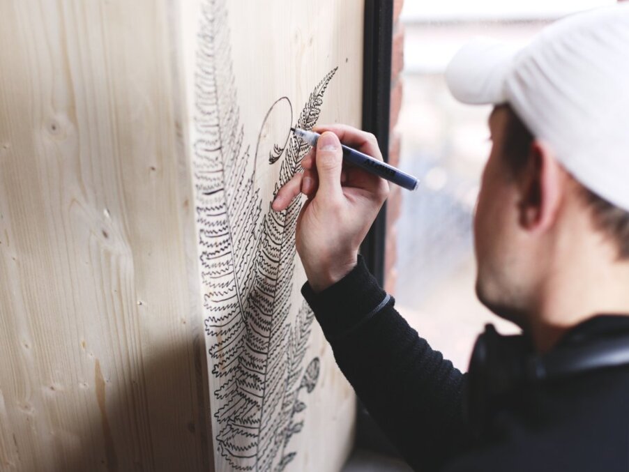 Artist drawing a detailed fern design with a pen on a wooden decorative panel