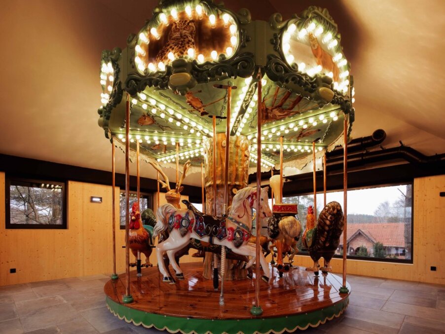 Indoor vintage carousel with hand-painted horses and animals, lit bulbs on polished wooden base in wood-paneled room with countryside view.