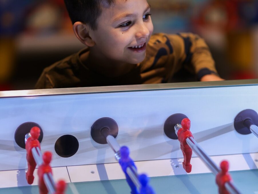 Smiling child playing foosball in hotel's brightly lit family game room, colorful table and casual play area.