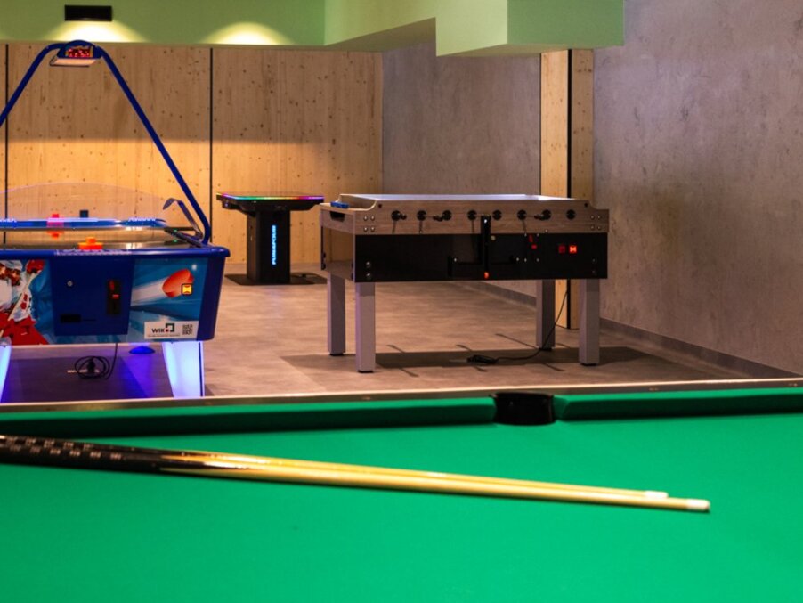 Hotel game room with green pool table and cues in foreground, air hockey and foosball tables, wood-paneled relaxed vibe.