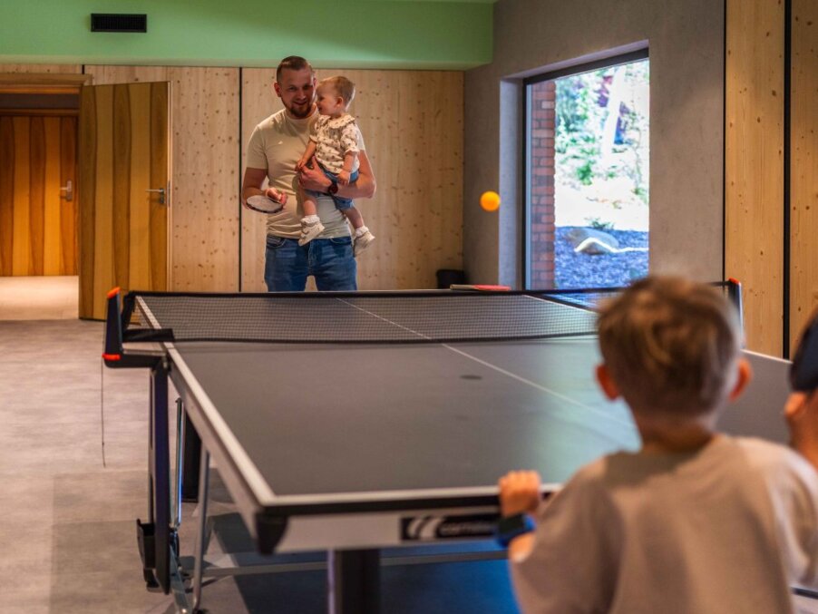 Family game room with indoor ping-pong table; father holding toddler while another child plays, warm wood paneling, garden view.
