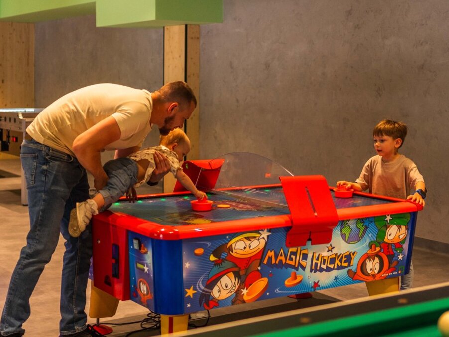 Hotel game room with colorful 'Magic Hockey' air-hockey table; father lifts toddler to play while older boy waits.