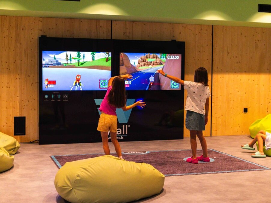 Children playing a motion-controlled racing game on a dual-screen gaming wall in a hotel kids' play area with beanbag seating.