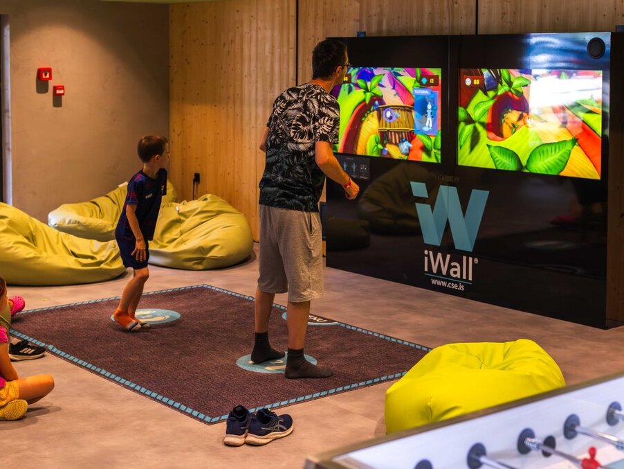 Family game room with interactive iWall dual screens, kids and adult playing on carpeted pads, lime bean bags, foosball table and warm wood paneling.