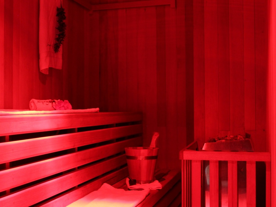Private wood-paneled sauna in red mood lighting, tiered bench with towels, wooden bucket and stone heater