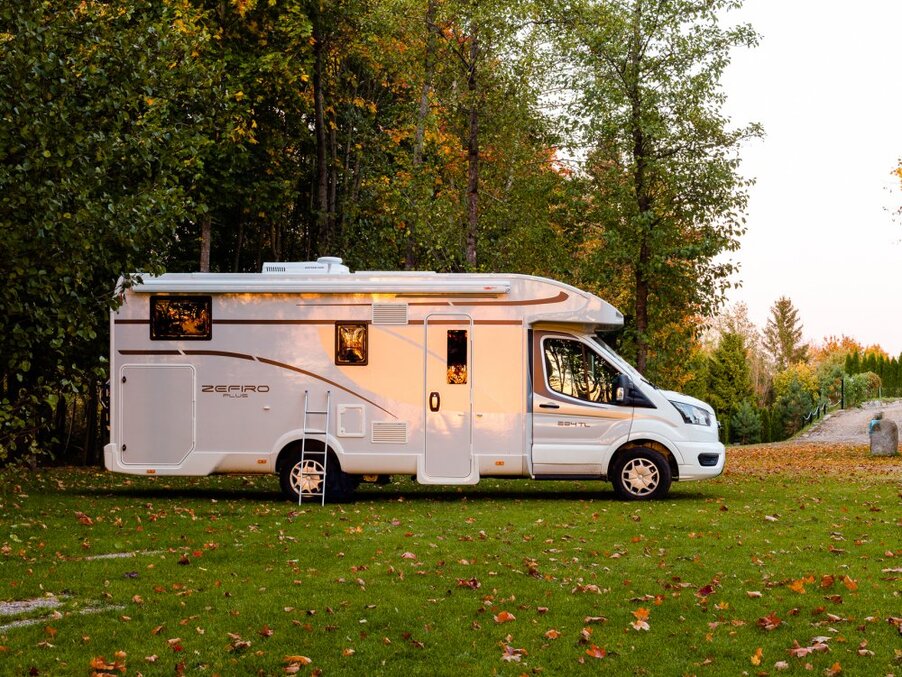 White motorhome parked on a grassy campsite by autumn trees; side door, roof vent and ladder visible, bathed in warm light