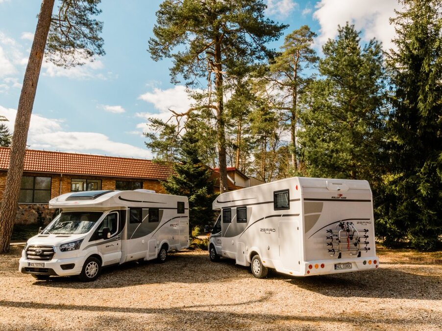 Two white motorhomes parked on a gravel lot beside a red-tiled lodge, bike rack on rear, surrounded by tall pines.