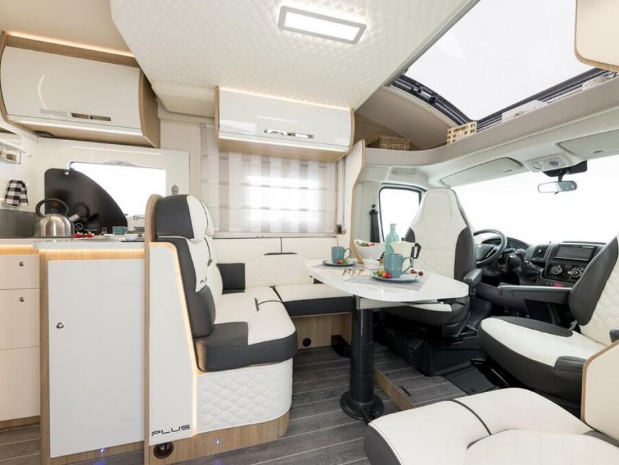 Modern motorhome interior with quilted white seating, dining table set for breakfast, compact kitchen and large skylight.