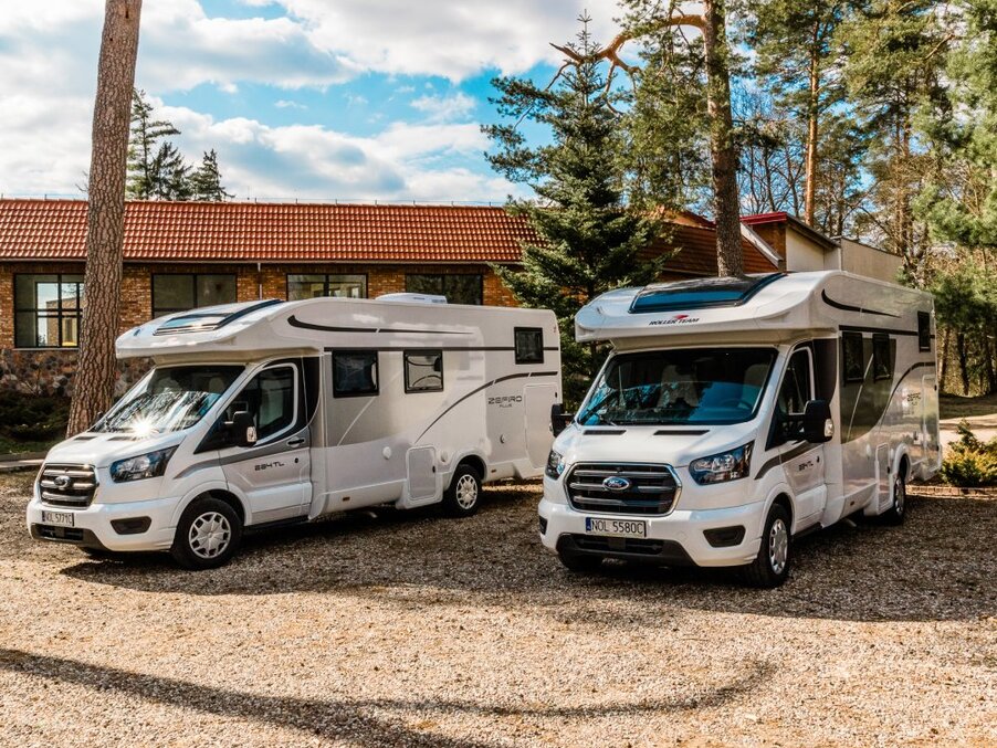 Two modern white motorhomes parked by a tiled-roof lodge amid pine trees, suggesting self-catering rentals for a peaceful forest getaway