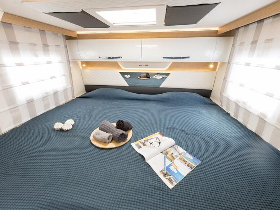 Cozy double cabin with skylight, large bed dressed in textured blue cover, overhead storage cabinets, sheer curtains, tray with towels and magazine.