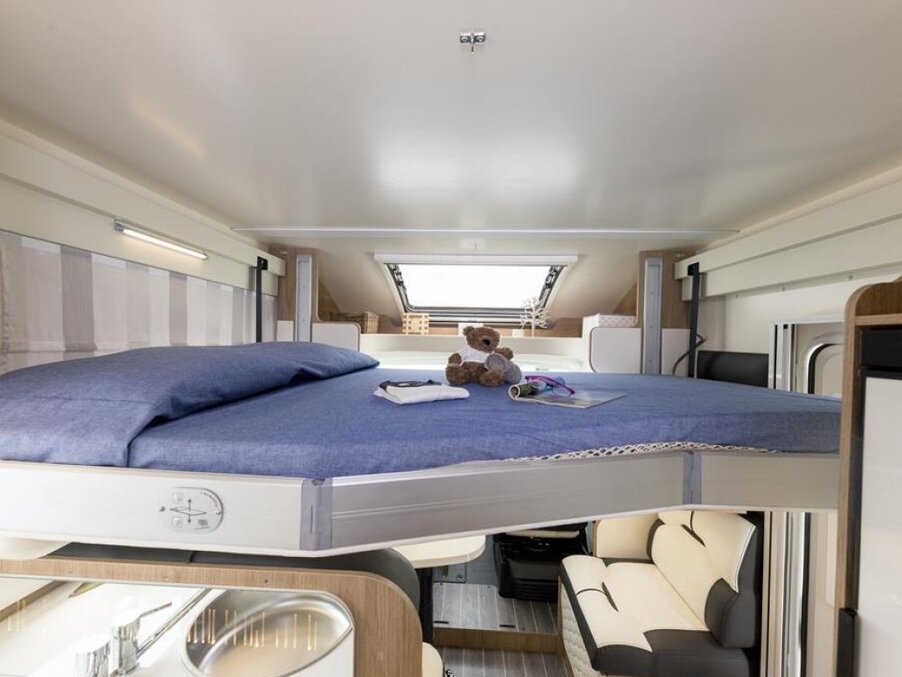 Over-cab double bed in a modern motorhome with blue linen, skylight, teddy, lounge seating and kitchenette below