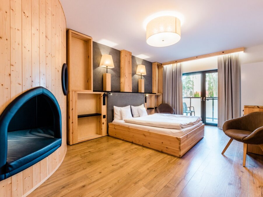 Warm wooden double room with low platform bed, bedside lamps, built-in cushioned nook, balcony with chairs and forest view.
