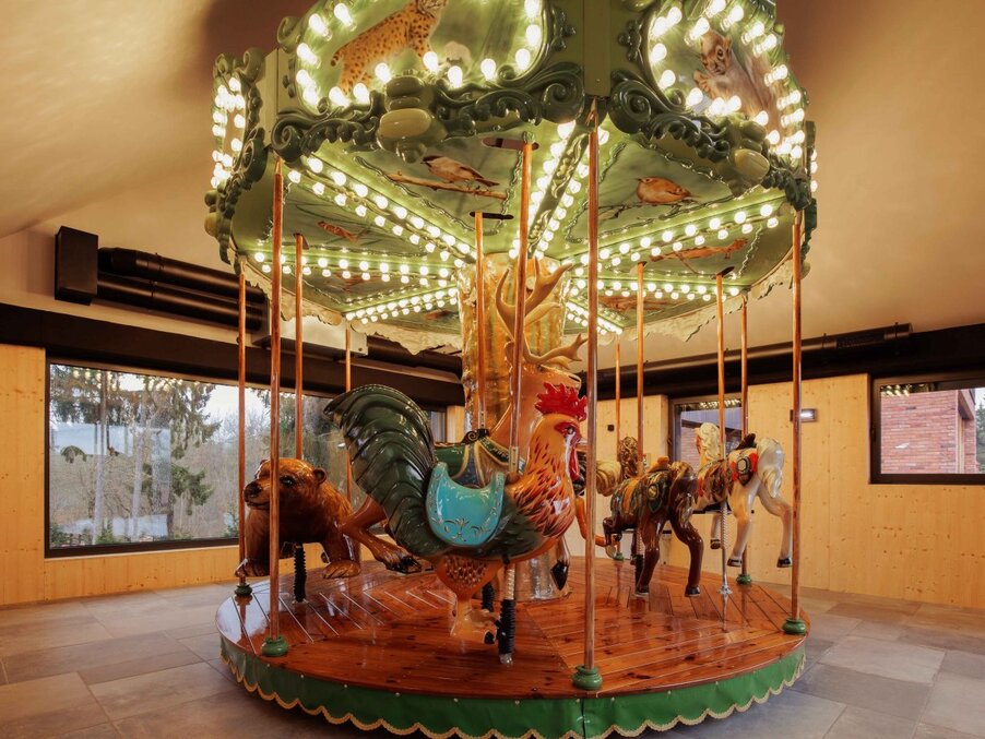 Warm-lit indoor vintage carousel with carved rooster, bear, horses and elephant on polished wooden platform in hotel family play area.