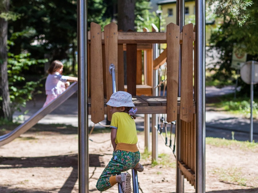 Wooden playhouse with metal slide rails in shaded pine grove; a child in a sun hat climbs while another uses the slide.