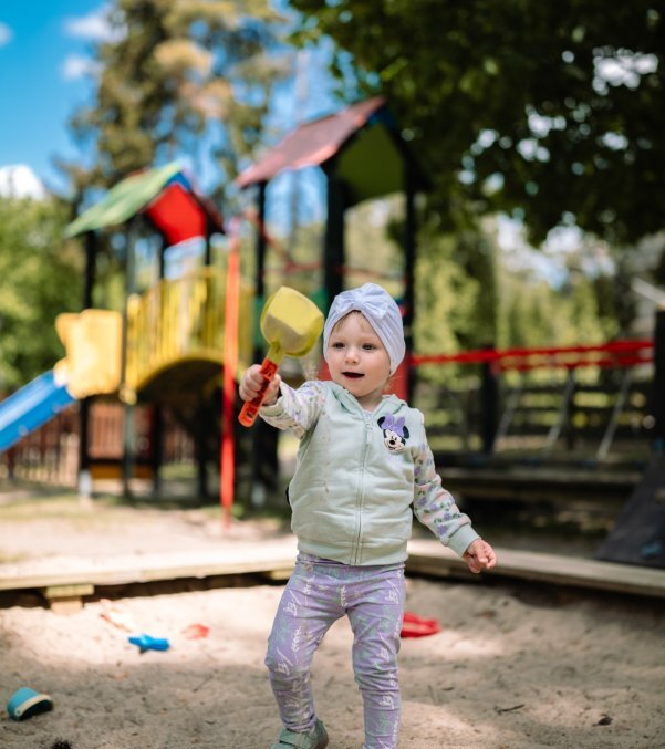 Toddler in sandbox holding a yellow shovel by a small sandcastle, colorful slides and climbing towers under trees in hotel family play area