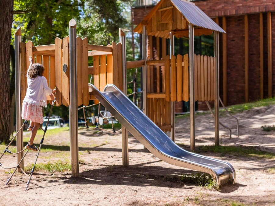 Family-friendly playground with wooden fort, metal slide and rope ladder as a child climbs under pine trees.