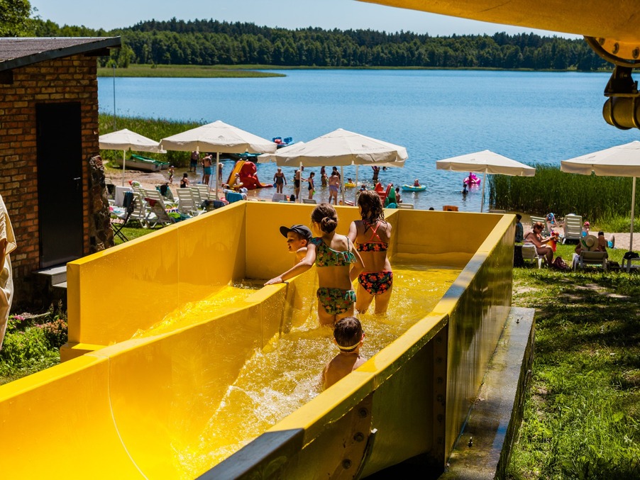 Children climbing a bright yellow water slide at a lakeside resort, sandy beach with umbrellas, sun loungers and swimmers