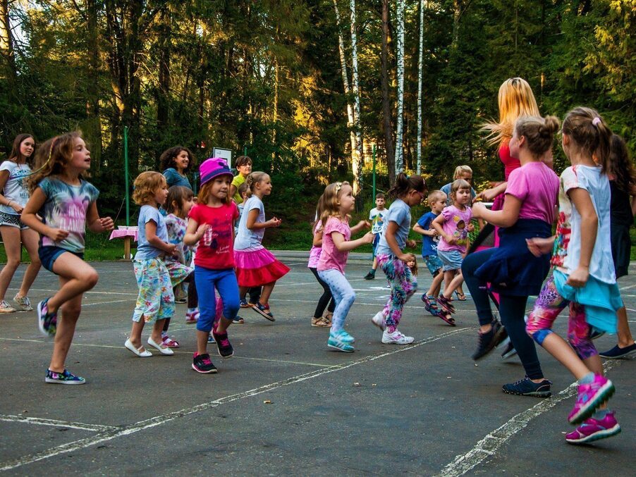 Group of children and an instructor jumping and dancing on an outdoor court surrounded by pine and birch trees