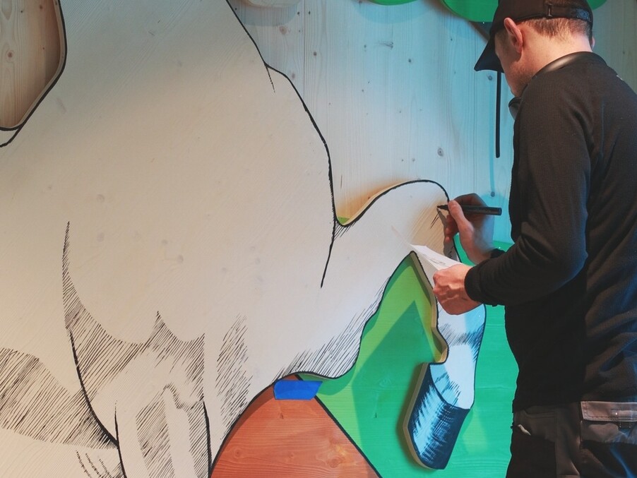 Artist sketching a large horse mural on wooden panels in a colorful interior, using a marker and reference sheet.