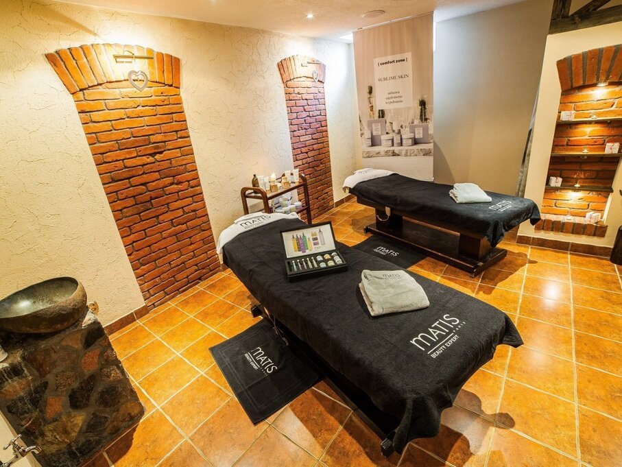 Couples spa treatment room with two massage tables, Matis towels, skincare display, warm tile floor and exposed brick arches.